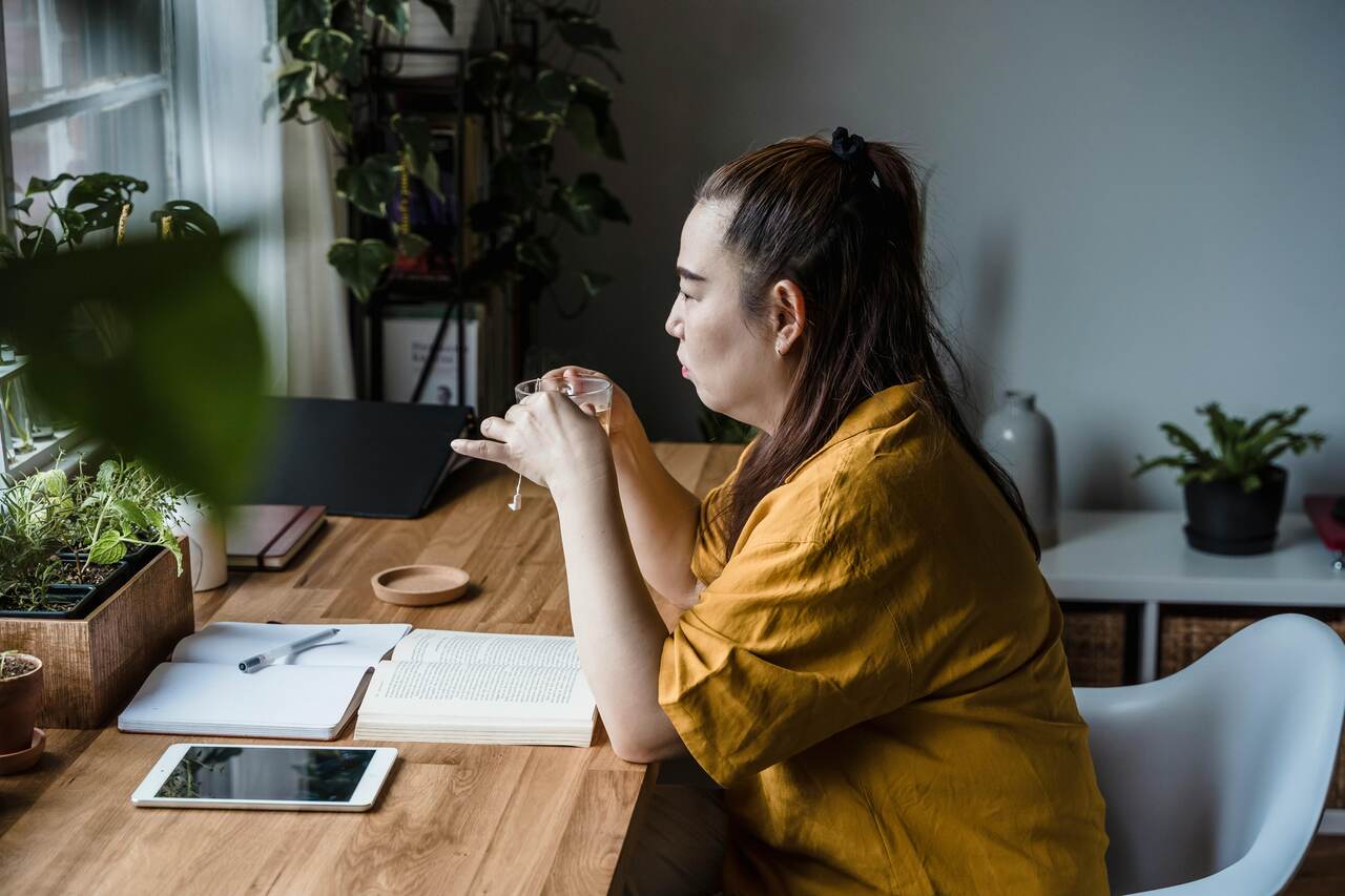 Mulher sentada à mesa com livros e tablet, tomando uma bebida quente enquanto faz anotações em um ambiente cheio de plantas.