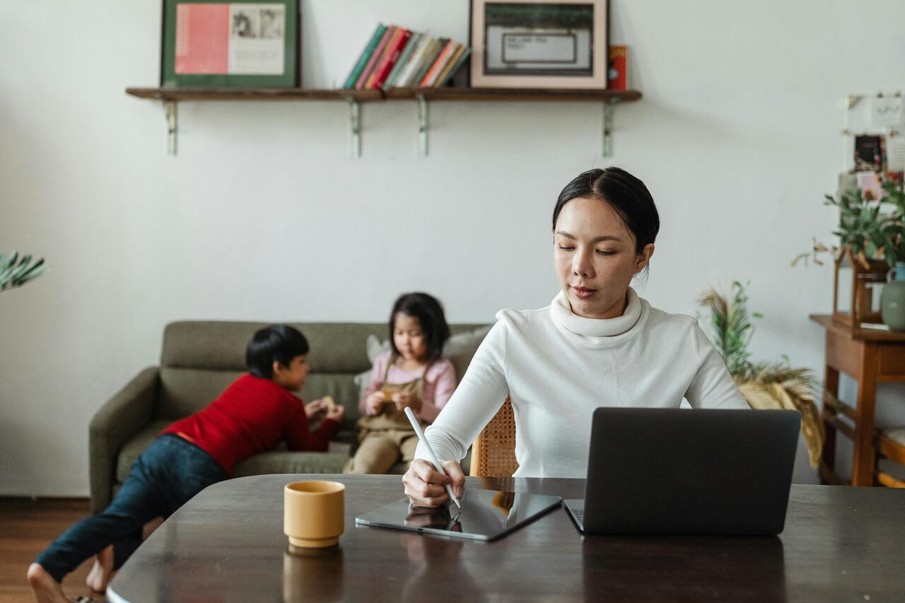 Mulher trabalhando em uma mesa com notebook e tablet, enquanto duas crianças brincam no sofá ao fundo em uma sala de estar.