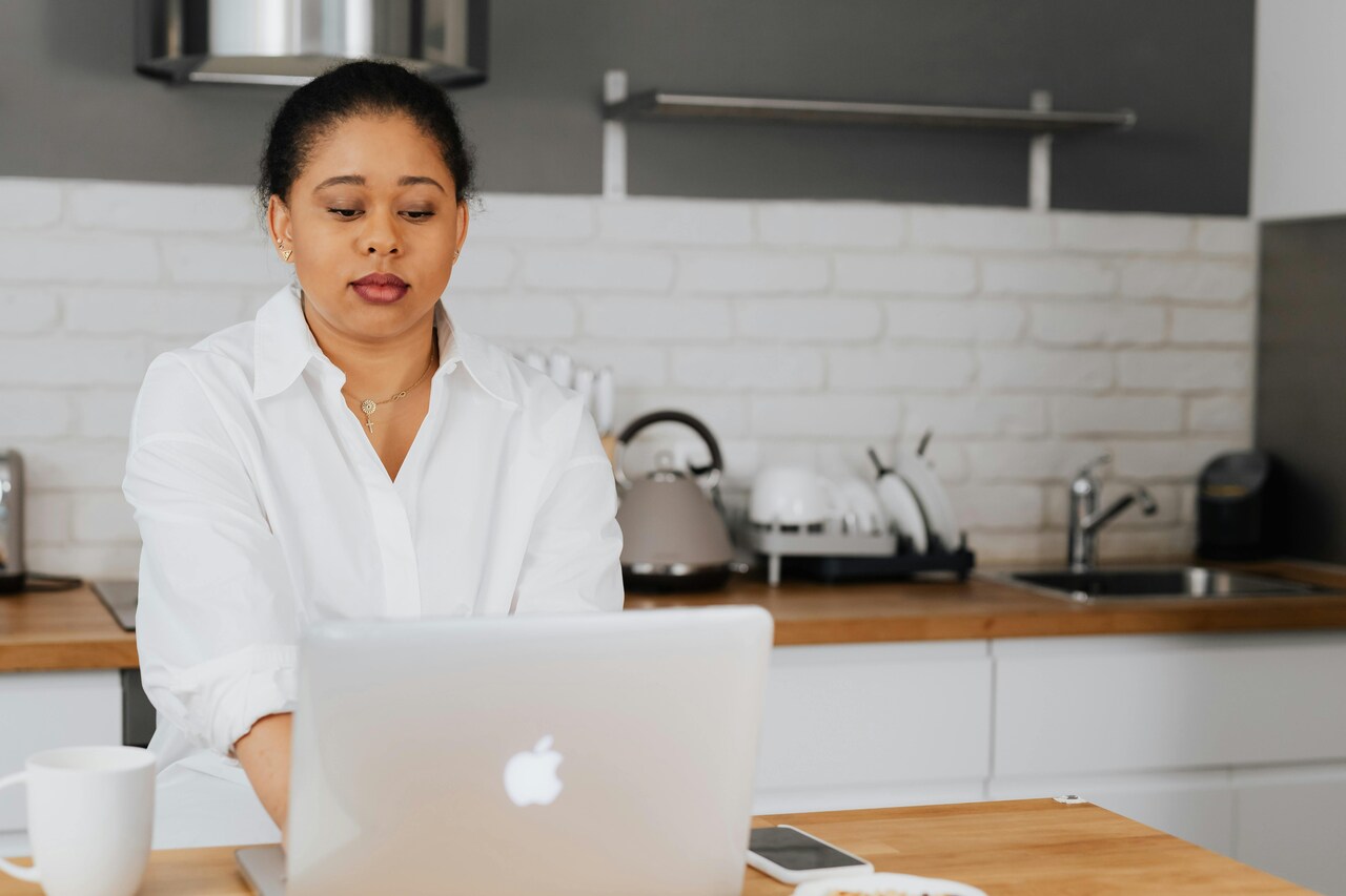 Mulher sentada na cozinha usando um notebook sobre a bancada, com expressão séria enquanto trabalha.