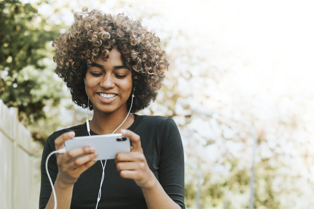 Uma mulher sorridente de cabelo cacheado está usando um celular enquanto passeia ao ar livre. Ela está sorrindo e segurando o celular, indicando a facilidade de fazer ajustes, como mudar o nome da rede Wi-Fi, diretamente pelo dispositivo. A imagem sugere a ideia de que modificar configurações simples, como o SSID da rede Wi-Fi, pode ser feito rapidamente em qualquer lugar.