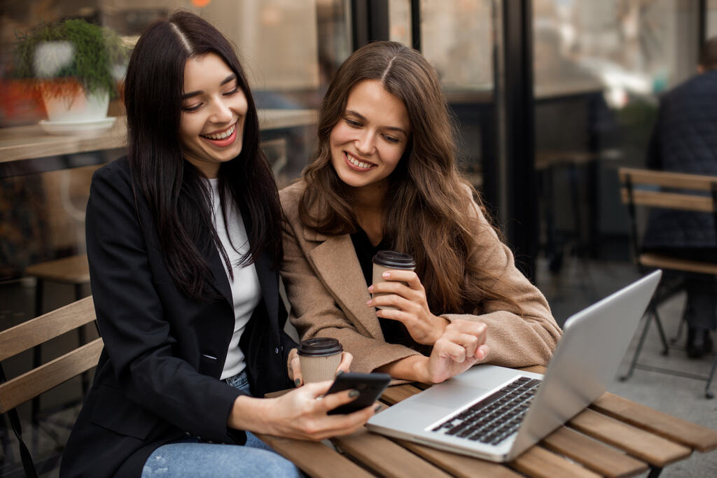 Duas mulheres sentadas em uma cafeteria, sorrindo enquanto olham para um celular, com um notebook e café sobre a mesa.