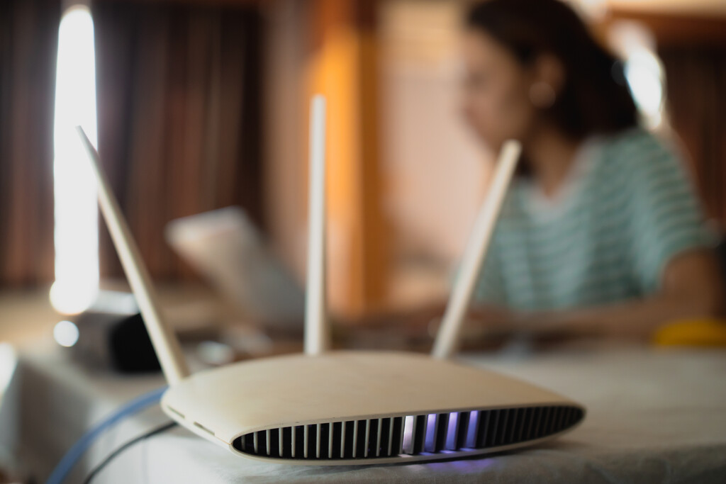 Roteador Wi-Fi ligado sobre a mesa com uma mulher usando notebook ao fundo, simbolizando conexão de internet banda larga.