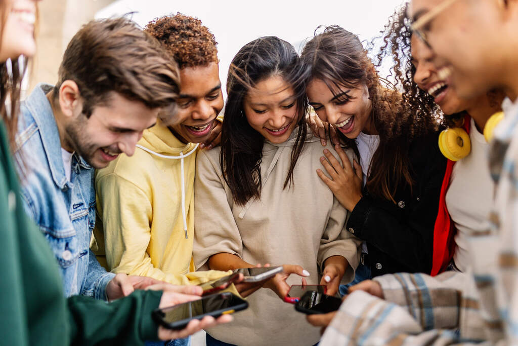Grupo de jovens reunidos ao ar livre, sorrindo e olhando para seus celulares enquanto compartilham algo divertido.