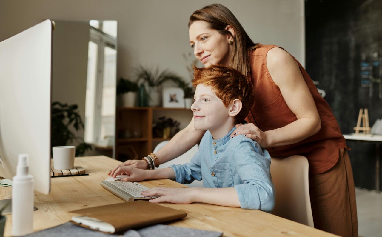 Mãe em pé atrás do filho, orientando enquanto ele usa um computador de mesa em um ambiente iluminado e organizado.