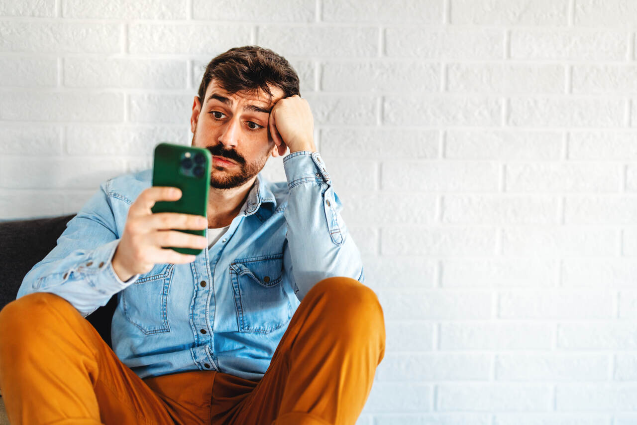 Homem sentado olhando para o celular com express&atilde;o de frustra&ccedil;&atilde;o, sugerindo problemas de conex&atilde;o ou internet lenta