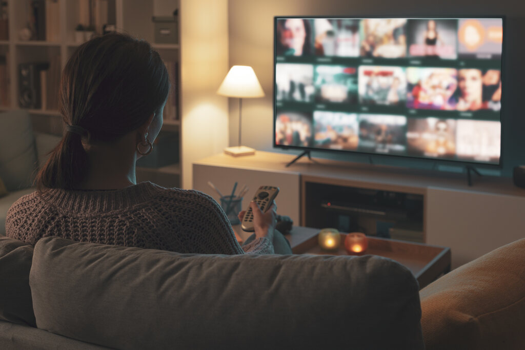 Mulher vista de costas sentada no sof&aacute; &agrave; noite escolhendo um filme no cat&aacute;logo da plataforma de streaming na TV.