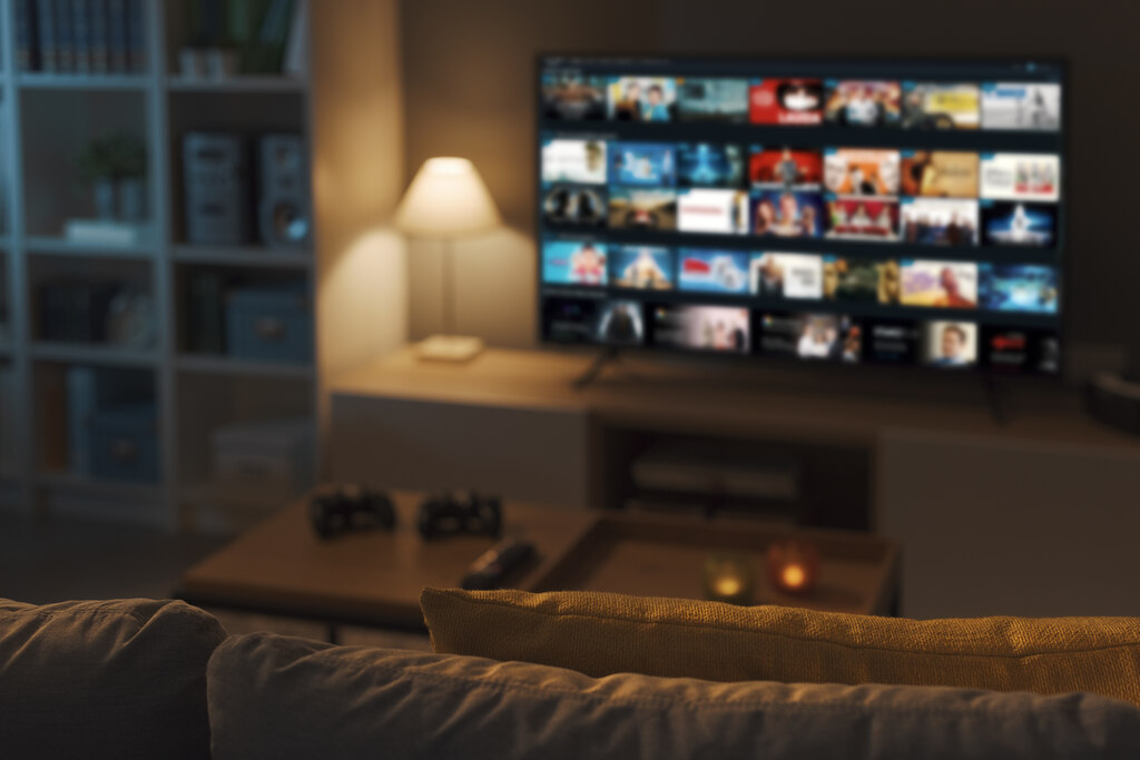 smart tv desfocada ao fundo exibindo cat&aacute;logo de filmes e s&eacute;ries em uma sala de estar moderna &agrave; noite.