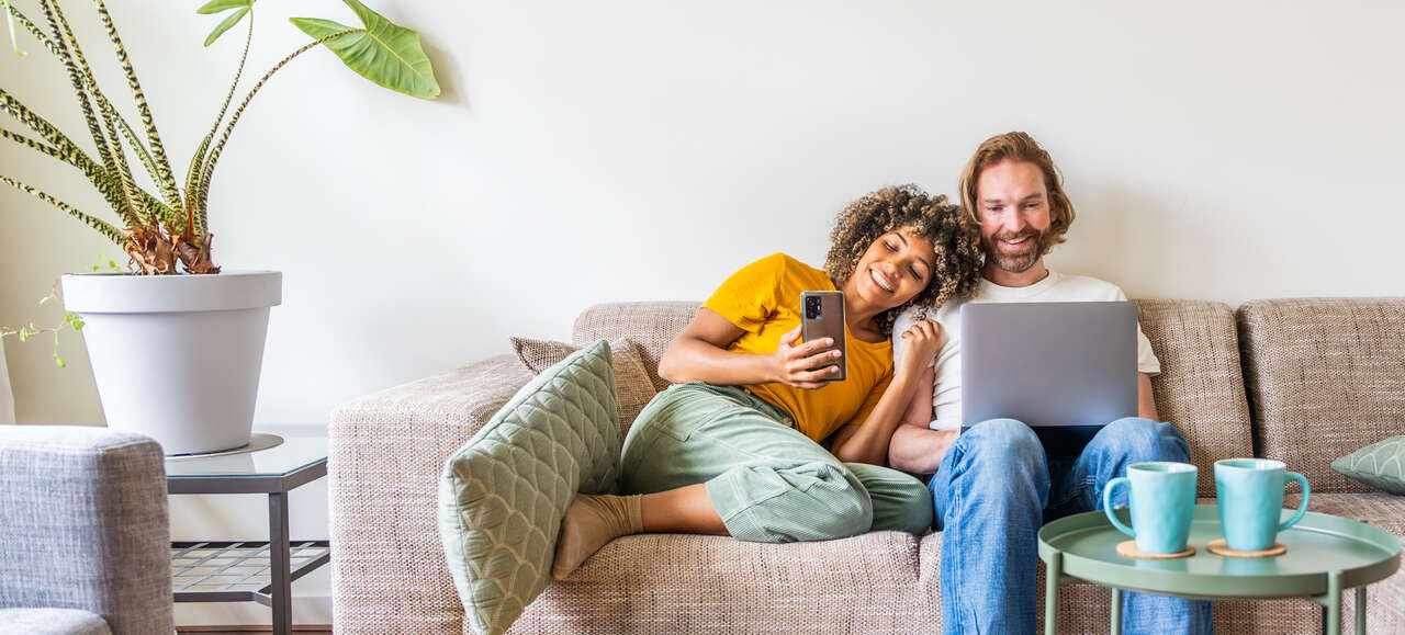 Casal sentado confortavelmente no sofá usando notebook e smartphone em casa, em ambiente aconchegante com decoração minimalista e planta ao lado.